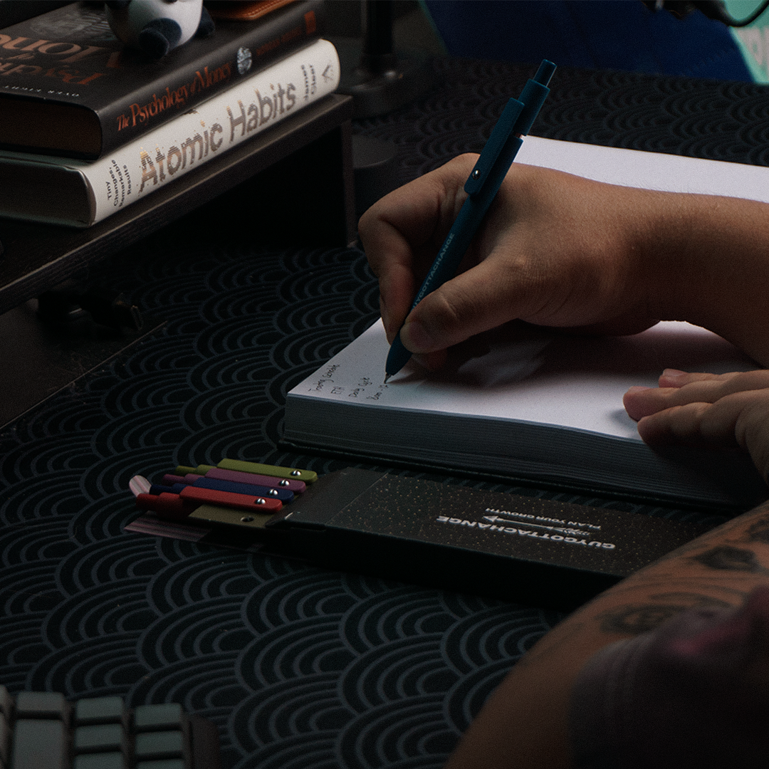 Writing notes in GuyGottaChange planner with black gel pen and 5-piece pen set box on desk