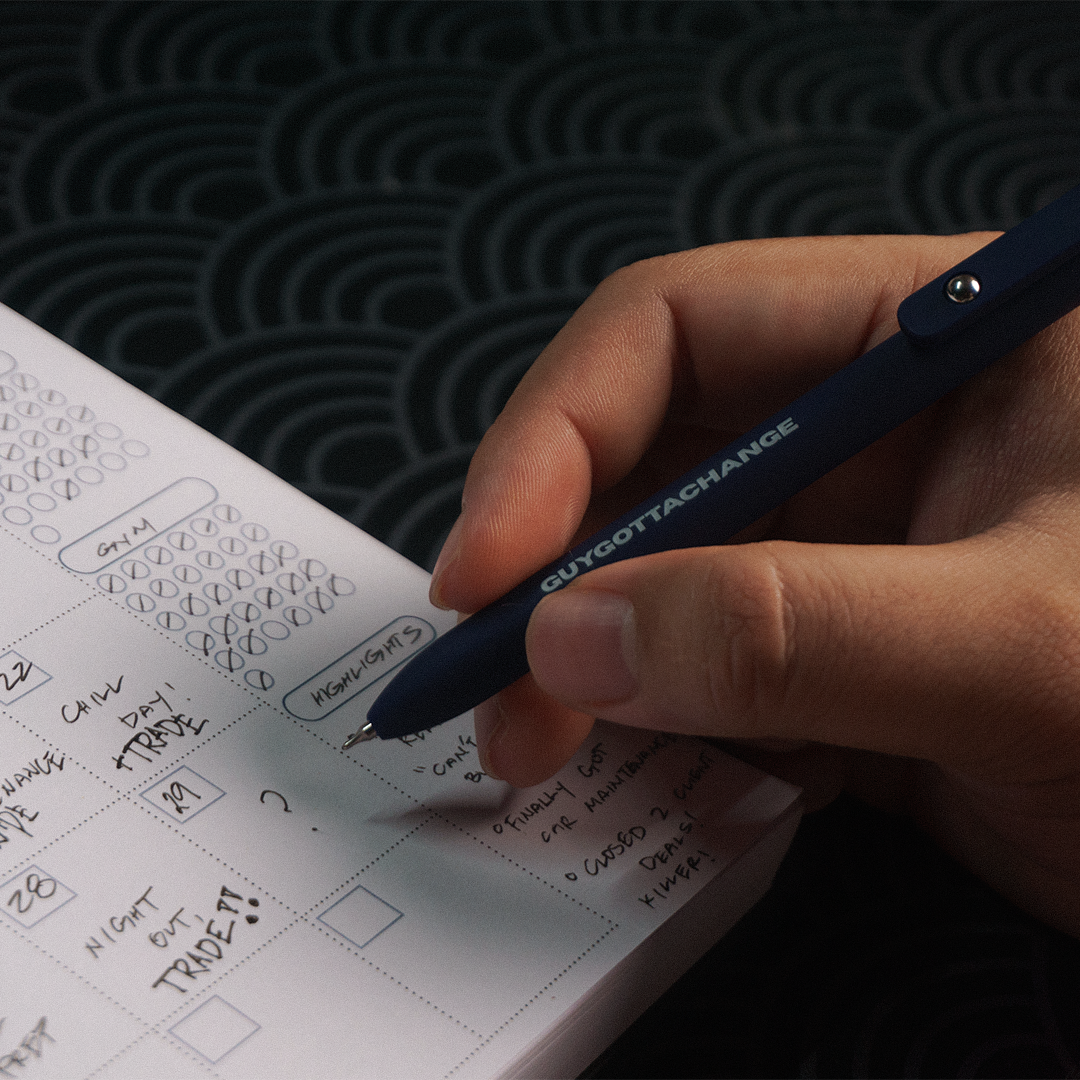 Close-up of GuyGottaChange branded black gel pen writing in weekly planner